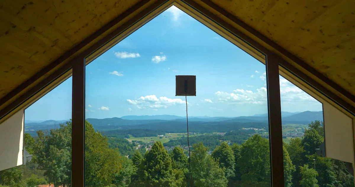 Panoramafenster mit Blick auf eine grüne Landschaft und Berge, in der Mitte ein Fensterputzroboter an der Scheibe befestigt.