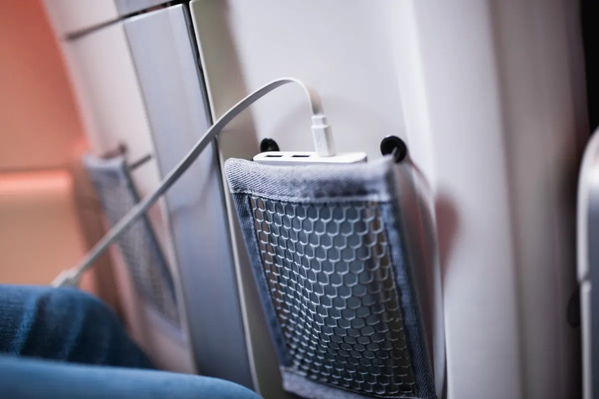 Una powerbank bianca ricarica un dispositivo nella tasca a rete di un sedile d’aereo.