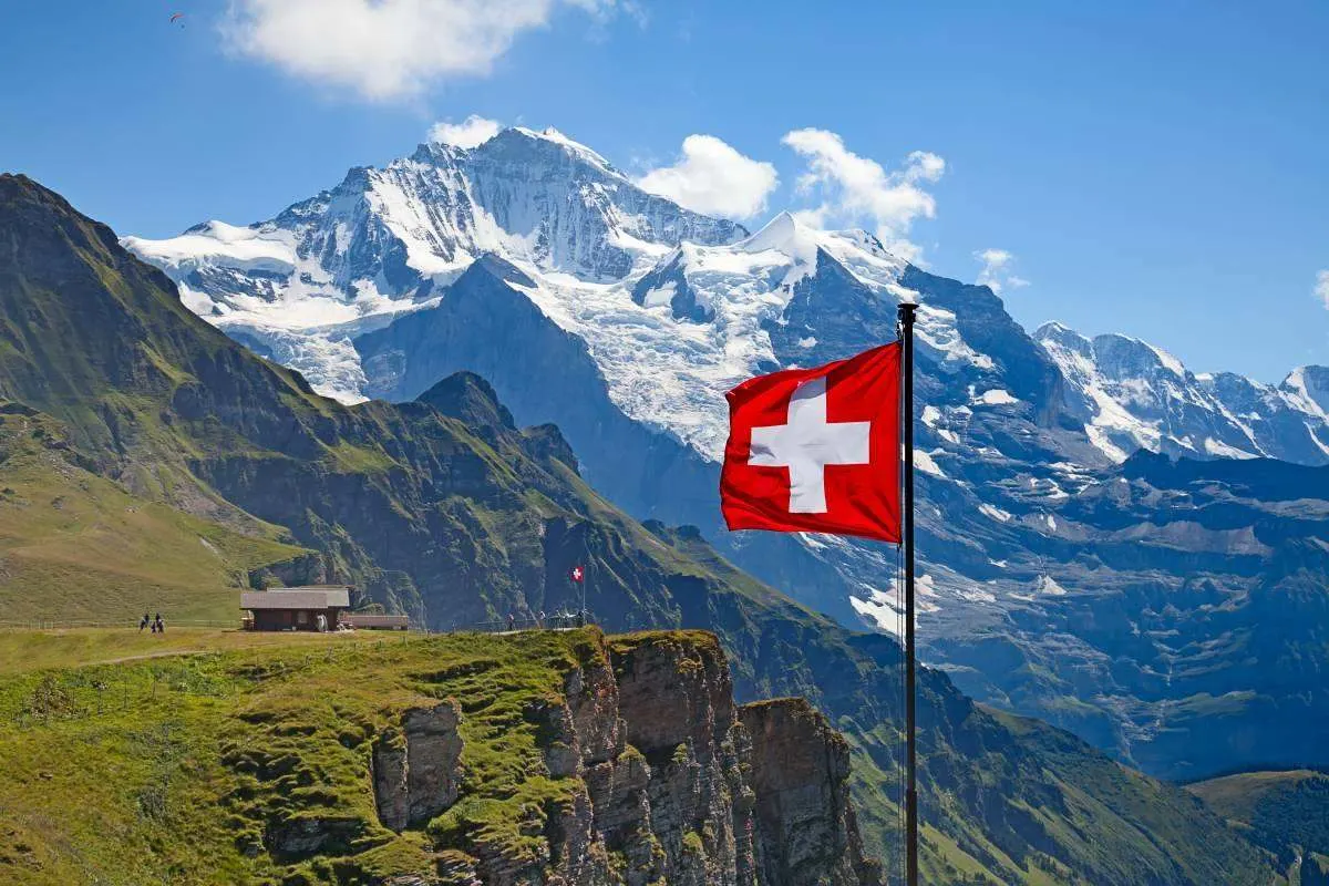Drapeau suisse devant des Alpes enneigées et un paysage verdoyant. 