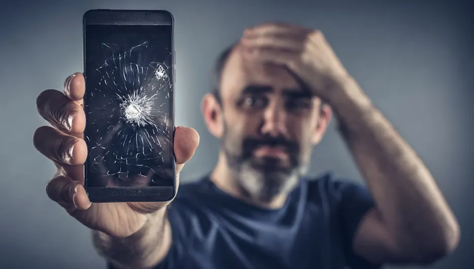 Un uomo tiene disperatamente in mano un natel con lo schermo incrinato davanti alla telecamera.