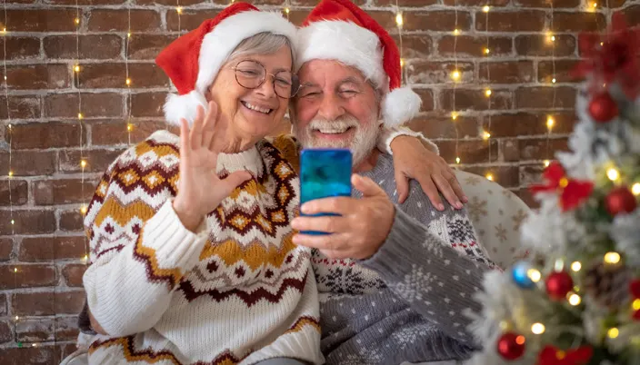 Grand-mère et grand-père avec des bonnets de Noël regardent un téléphone portable, avec un sapin de Noël au premier plan.