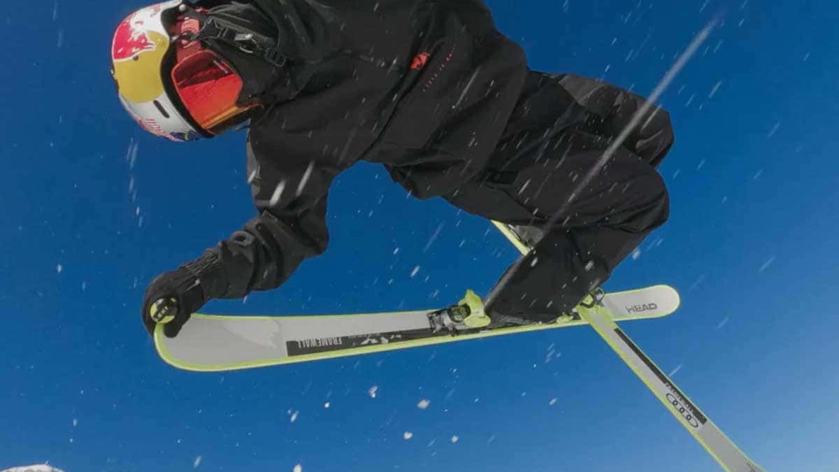 Un skieur en tenue noire et casque rouge saute sur des skis blancs contre un ciel bleu clair.