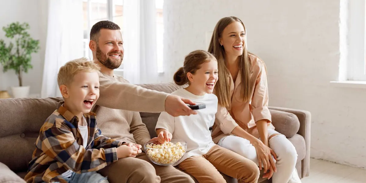 Eine Familie sitzt auf einem Sofa und lacht. Der Vater hält eine Fernbedienung, während die Kinder Popcorn essen.