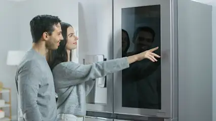 Ein Mann und eine Frau stehen vor einem Side by Side Kühlschrank, Der Kühlschrank ist silberfarben und hat ein integriertes Display