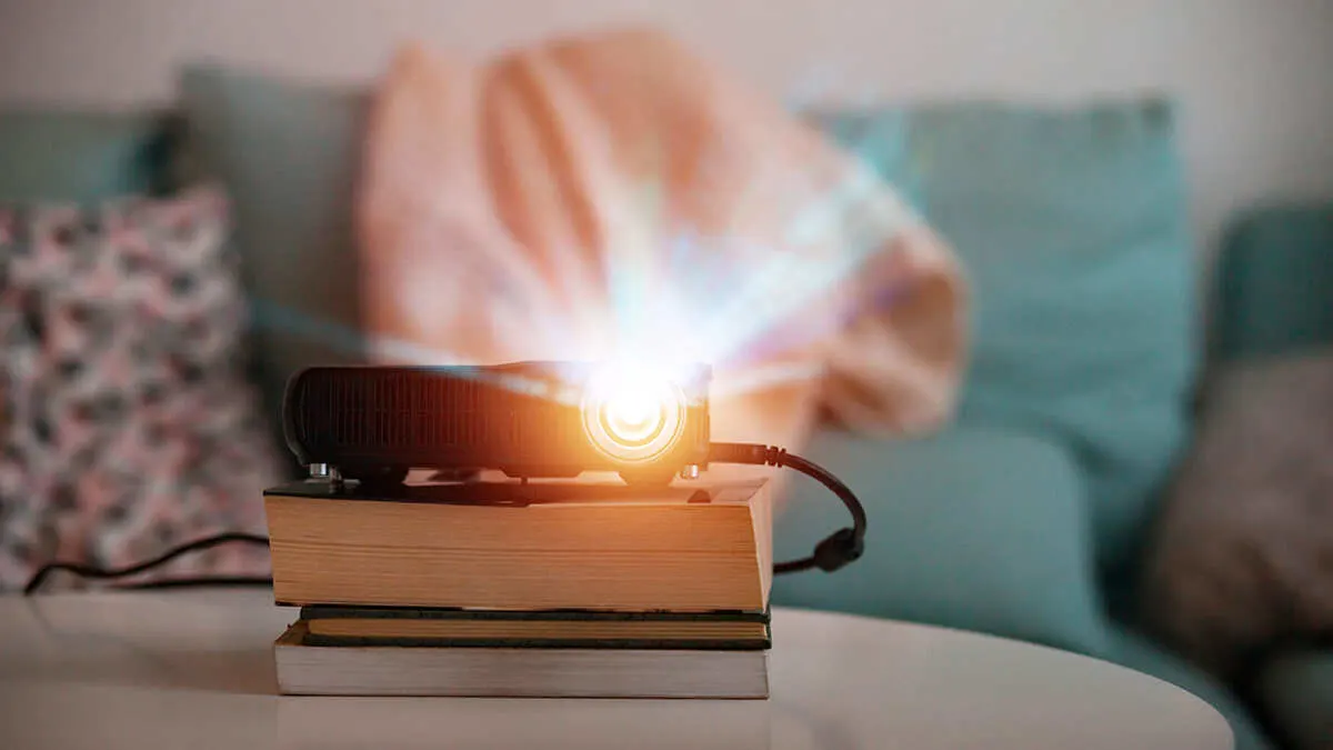 Ein Projektor steht auf einem Bücherstapel und wirft helles Licht. Im Hintergrund sind Kissen und ein Sofa zu sehen.