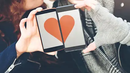 Deux personnes tiennent des téléphones affichant un cœur orange sur fond blanc. Les vêtements sont noirs et à carreaux.