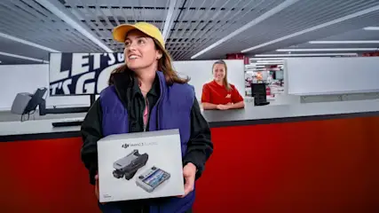 Frau hält DJI Mavic 3 Classic in einem Geschäft mit roter Theke und Kassiererin im Hintergrund.