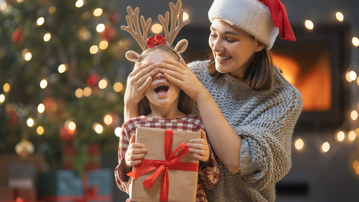 Una madre con cappello da Babbo Natale benda gli occhi a una bambina con cerchietto da renna. La bambina tiene un regalo con fiocco rosso.