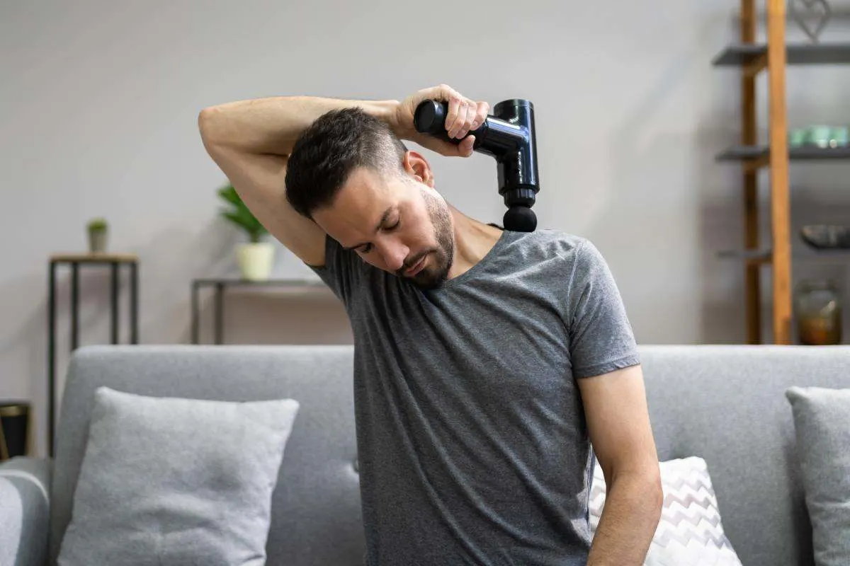 Homme assis sur un canapé massant sa nuque et ses épaules avec un pistolet de massage. 