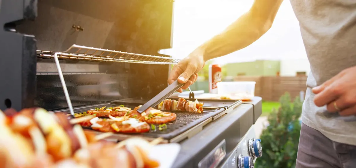 Persona che gira carne e spiedini su un barbecue a gas nella calda luce della sera. 