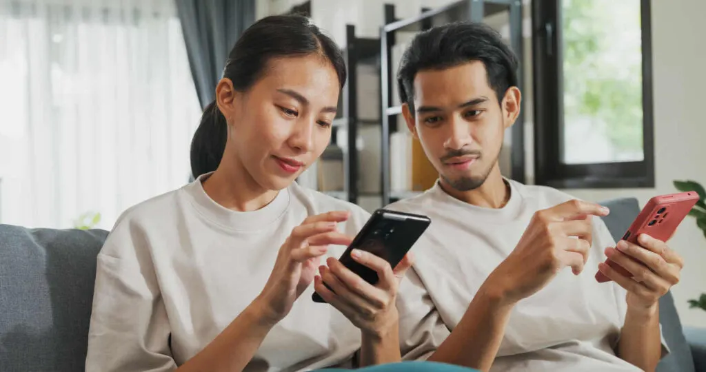 Una giovane coppia è seduta sul divano e tiene ciascuno uno smartphone in mano. 