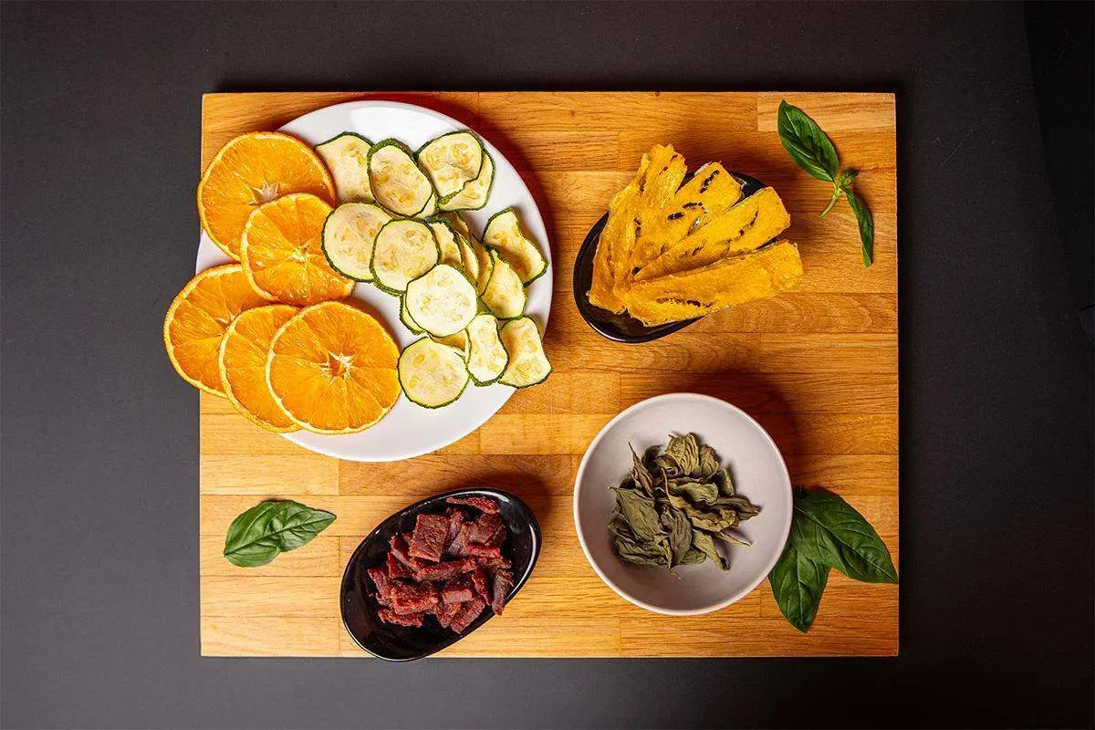 Tranches d’oranges, de courgettes et de légumes séchés, accompagnées d’autres snacks sur une planche en bois.
