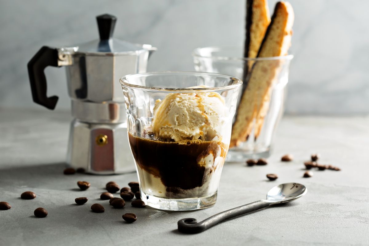 Gelato alla vaniglia con caffè in un bicchiere, moka argentata, cucchiaio, chicchi di caffè e biscotti su superficie grigia.