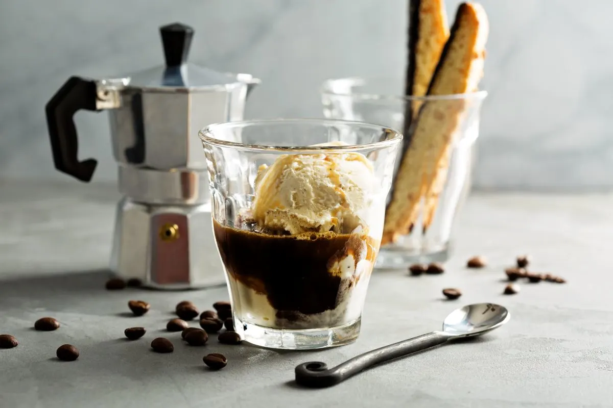 Affogato mit Vanilleeis und Kaffee in Glas, Kaffeebohnen, Löffel, Mokkakocher und Biscotti auf grauem Tisch.