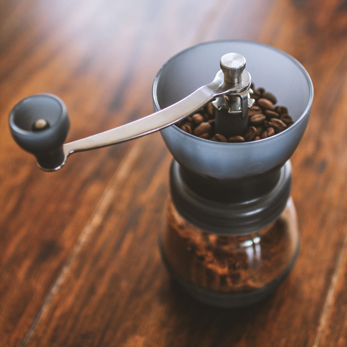Dunkelgraue manuelle Kaffeemühle mit silberner Kurbel auf braunem Holztisch. Oben Kaffeebohnen, unten gemahlener Kaffee.