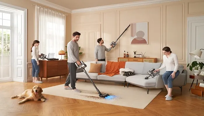 Quatre personnes utilisent des aspirateurs dans un salon beige avec un chien couché sur le sol.