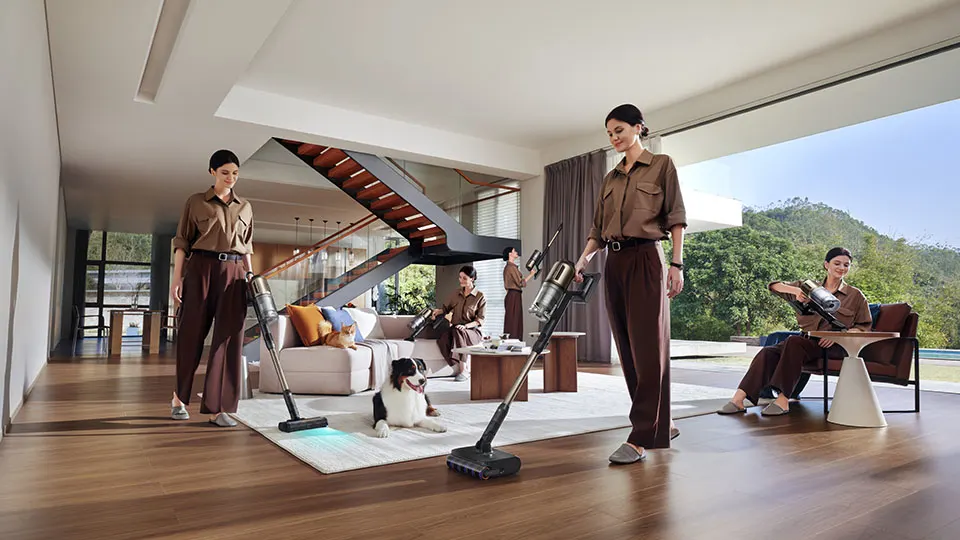 Quatre personnes utilisent des aspirateurs dans un salon beige avec un chien couché sur le sol.