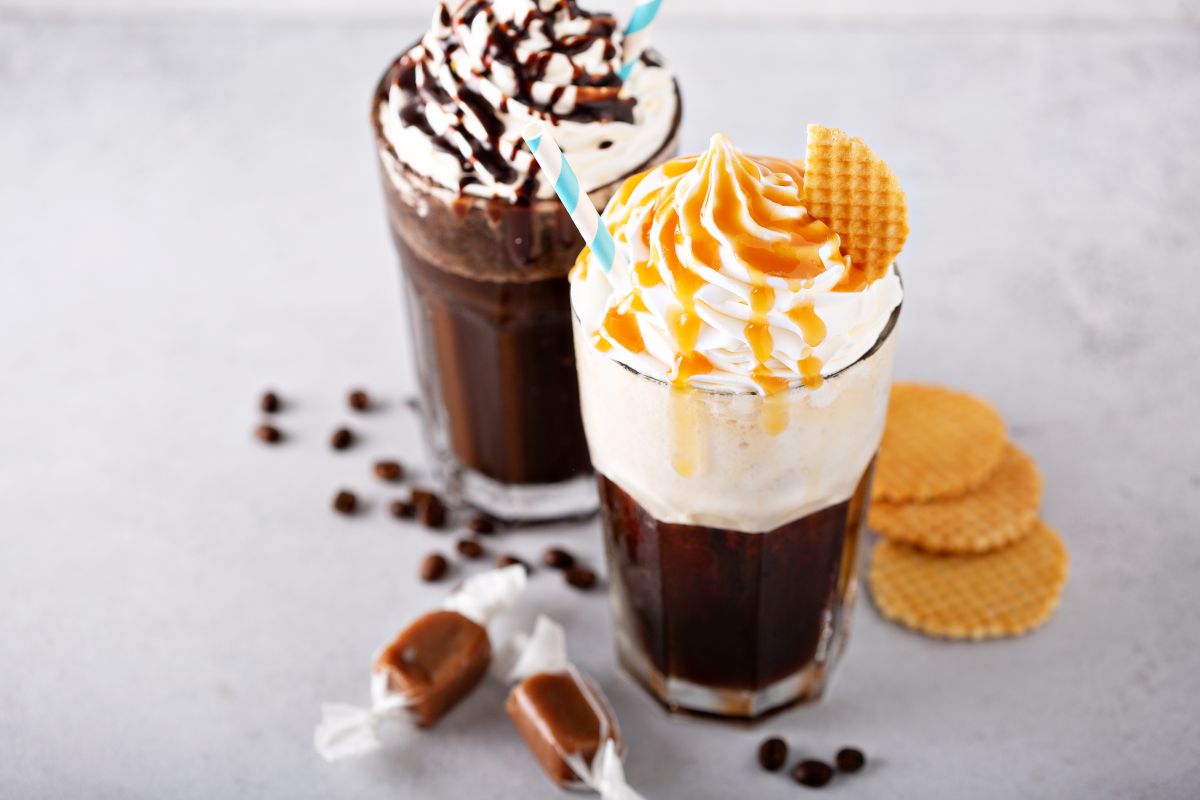 Zwei Eiskaffee-Gläser mit Sahne, blau-weiss gestreiften Strohhalmen. Eines mit Karamell, das andere mit Schokolade. Dazu Waffeln, Kaffeebohnen, Karamellbonbons auf grauem Untergrund.