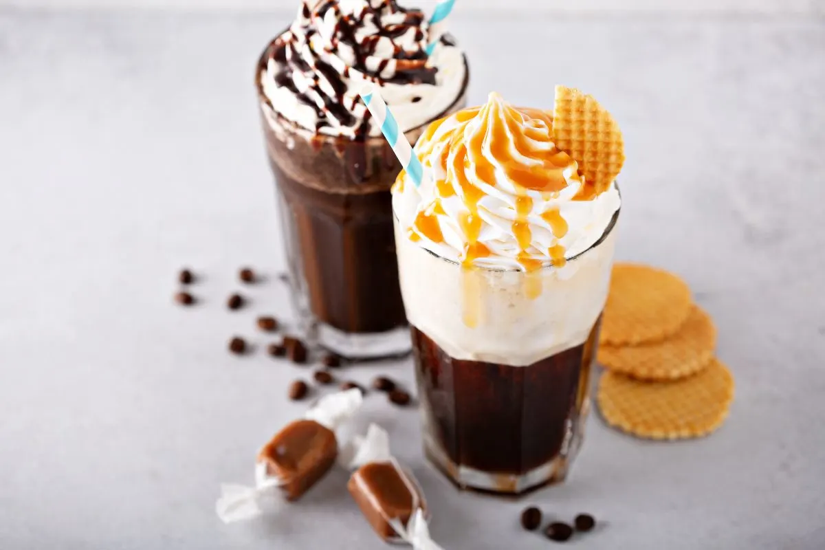 Zwei Eiskaffee-Gläser mit Sahne, blau-weiss gestreiften Strohhalmen. Eines mit Karamell, das andere mit Schokolade. Dazu Waffeln, Kaffeebohnen, Karamellbonbons auf grauem Untergrund.