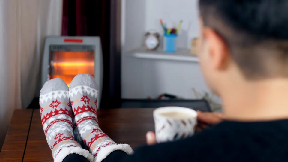 Un homme portant des chaussettes de Noël à motifs rouges et gris boit un café, avec un radiateur mobile en arrière-plan.