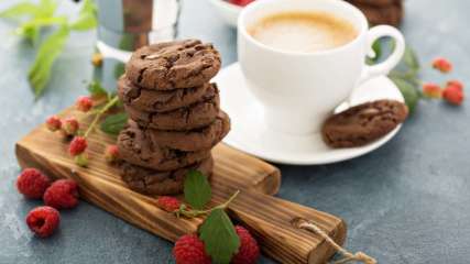 Ein Stapel Schoko-Cookies auf einem Holzbrett, garniert mit Himbeeren und eine Tasse Kaffee im Hintergrund.
