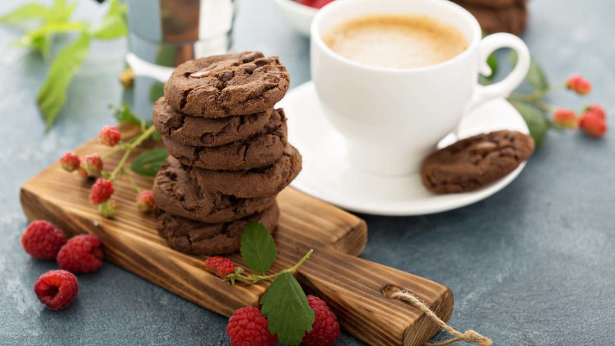 Tazza bianca con caffè, biscotti al cioccolato su tagliere di legno e lamponi rossi.