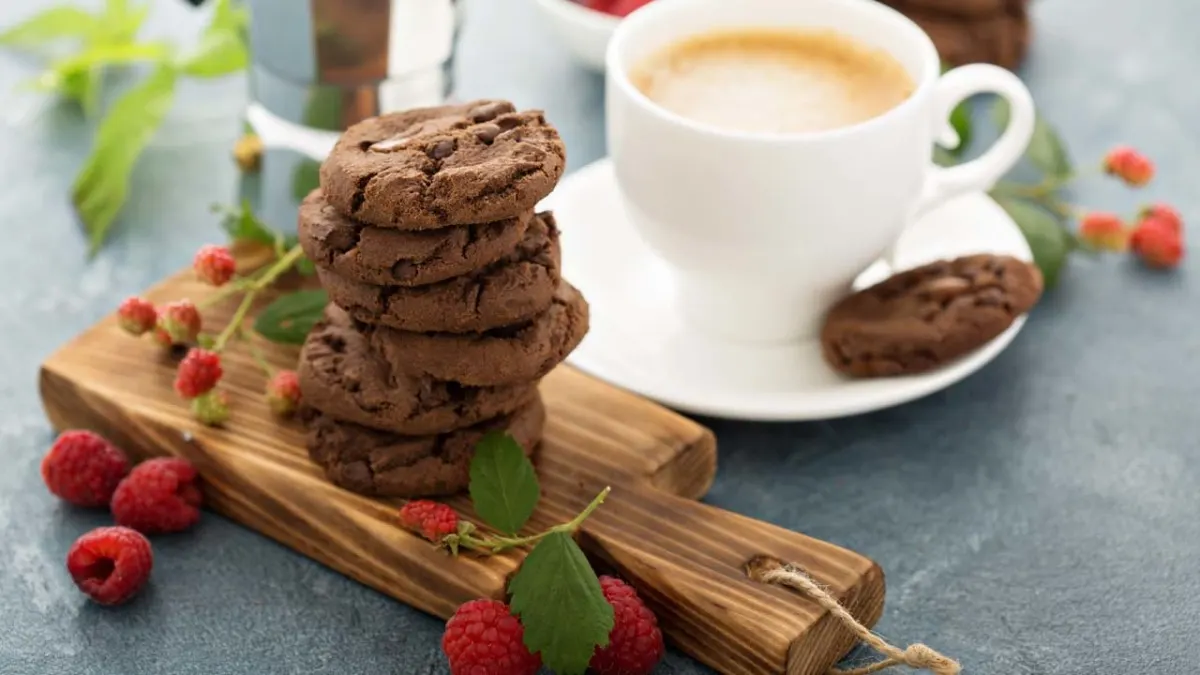 Tazza bianca con caffè, biscotti al cioccolato su tagliere di legno e lamponi rossi.