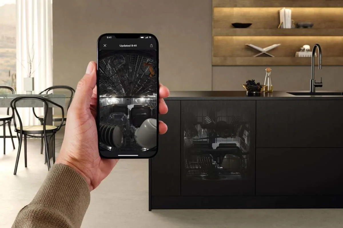 Una persona tiene uno smartphone davanti a una lavastoviglie Miele in una cucina moderna. Sul display si vede l’interno della lavastoviglie con le stoviglie caricate.