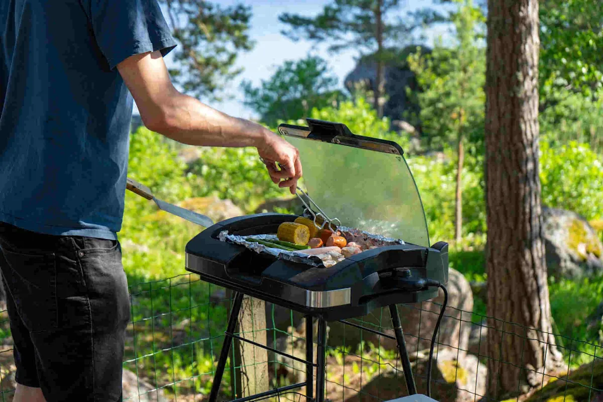 Une personne fait des grillades sur un barbecue électrique ouvert dans le jardin, sur lequel se trouvent des épis de maïs, des légumes et de la viande.
