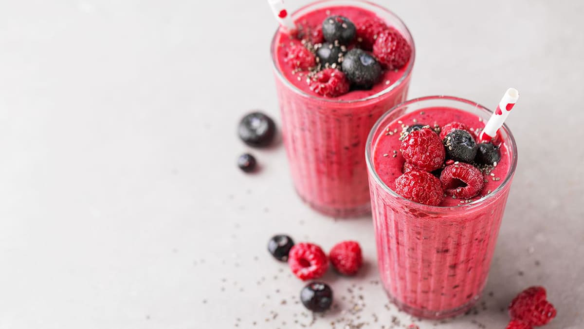 Deux verres de smoothie rose garnis de framboises et de myrtilles avec des pailles sur une surface grise.