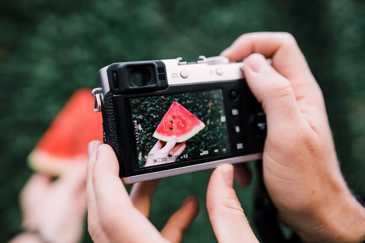 Mani che tengono una fotocamera compatta e fotografano un pezzo di anguria visibile sul display.