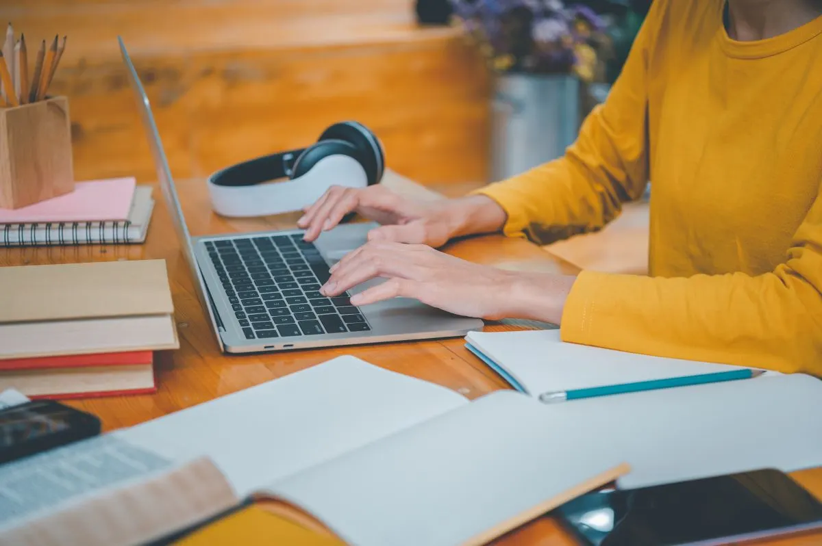 Détail d'un bureau encombré de livres et de cahiers, une personne travaillant sur un ordinateur portable