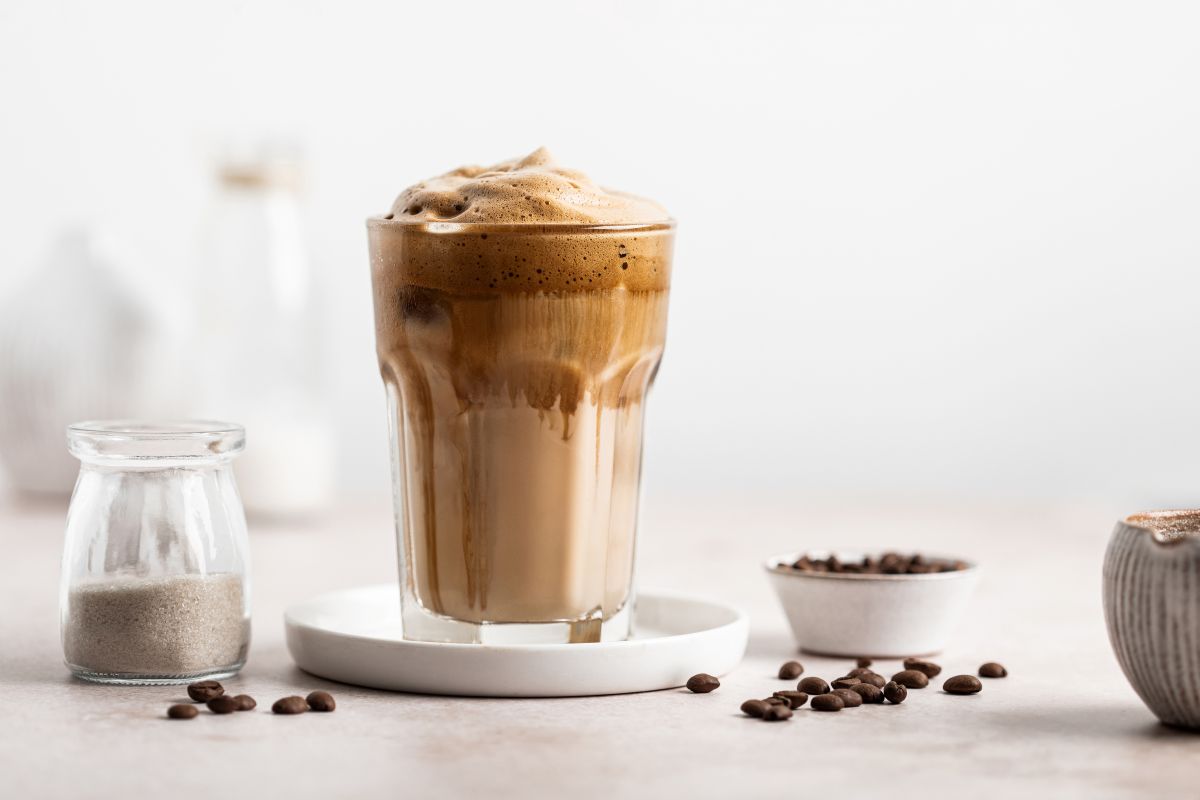 Eisgekühlter Kaffee mit dunklem Schaum in hohem Glas auf weisser Untertasse. Daneben Zuckerglas, Kaffeebohnen, Schale und Topf vor hellem Hintergrund.