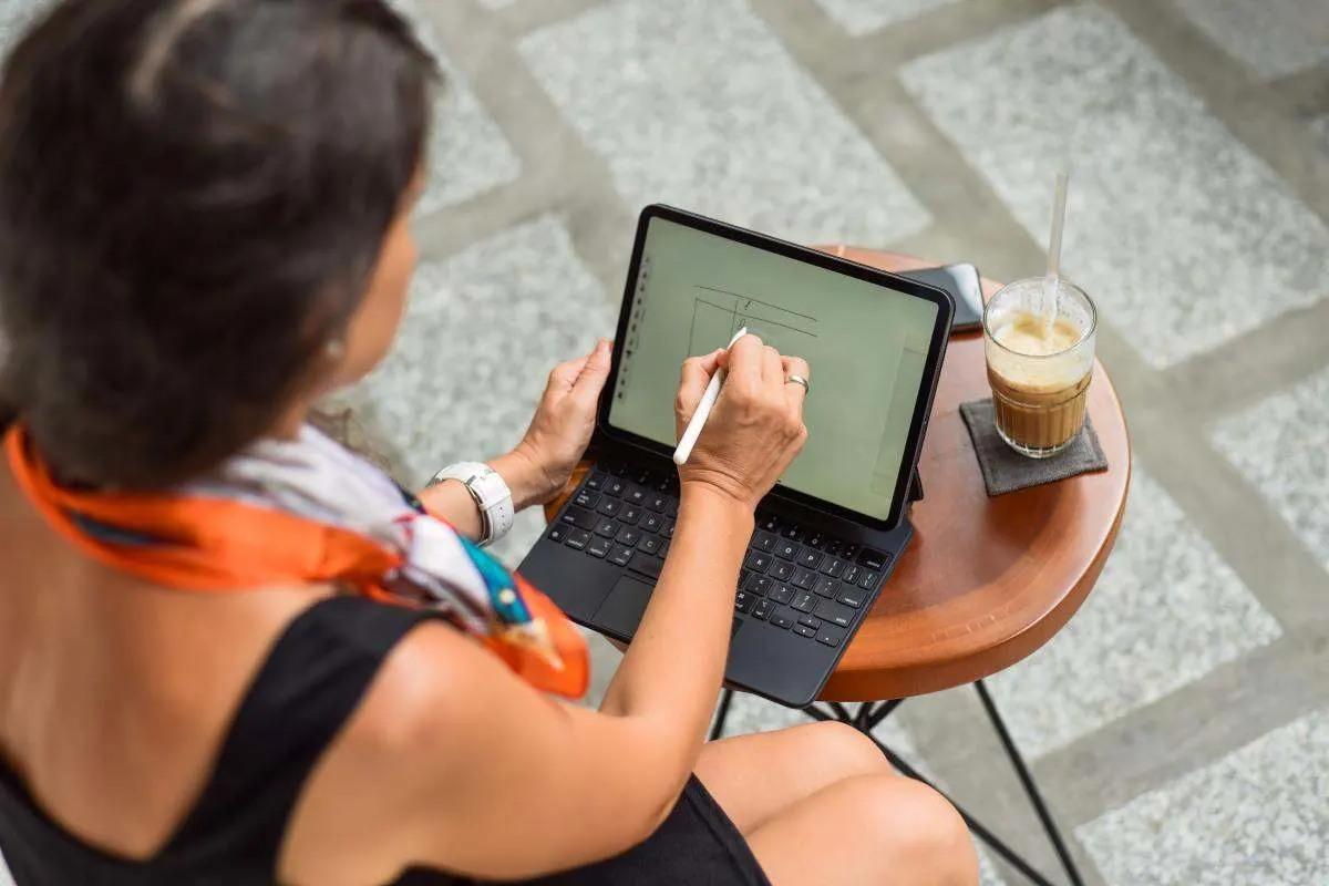 Une femme écrit avec un stylet sur une tablette avec clavier à une table de café.