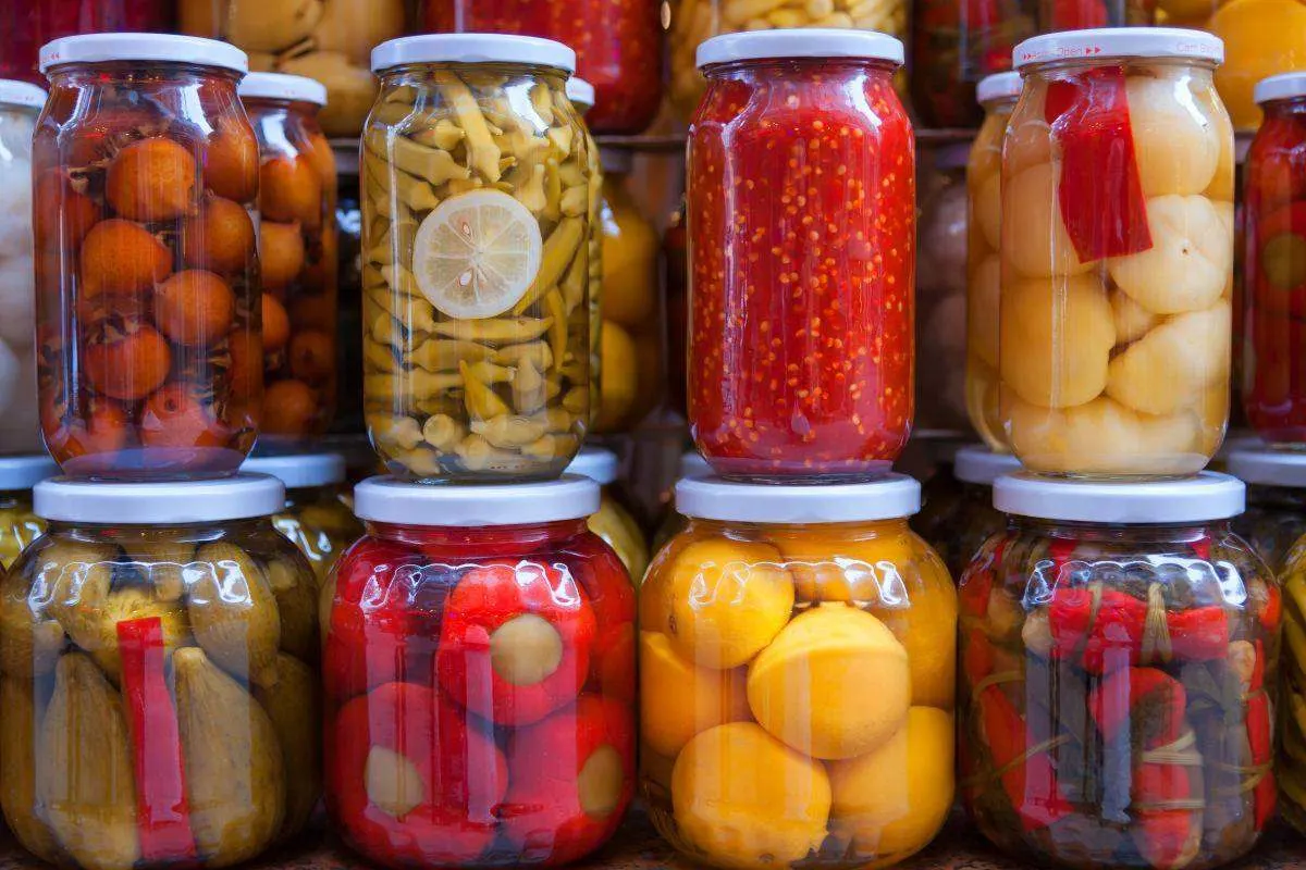 Varie verdure e frutti in conserva in barattoli di vetro, disposti fittamente uno accanto all'altro in più file.