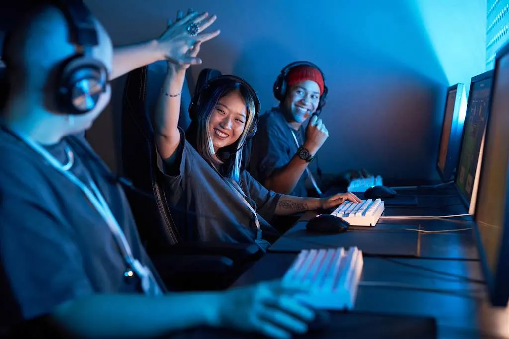 Tre gamer con cuffie sono seduti uno accanto all’altro davanti a PC da gaming. Al centro, una giovane donna sorridente alza la mano per dare il cinque mentre il gruppo gioca insieme.