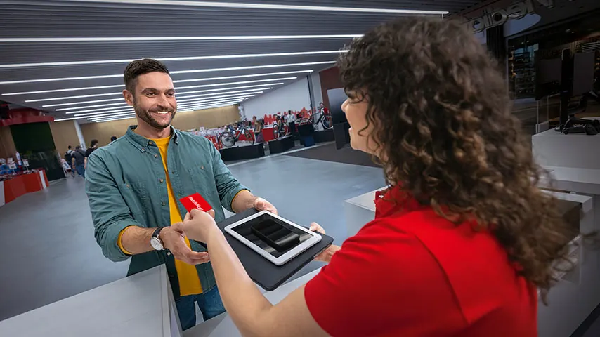 Un homme donne une carte rouge à une femme tenant une tablette noire. L'arrière-plan montre un magasin bien éclairé avec des présentoirs.