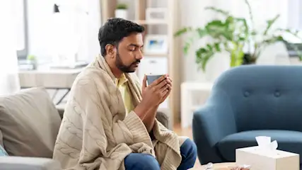 Mann sitzt auf dem Sofa im Wohnzimmer, in eine Decke eingekuschelt, wärmt seine Hände an einer Tasse mit heissem Getränk.