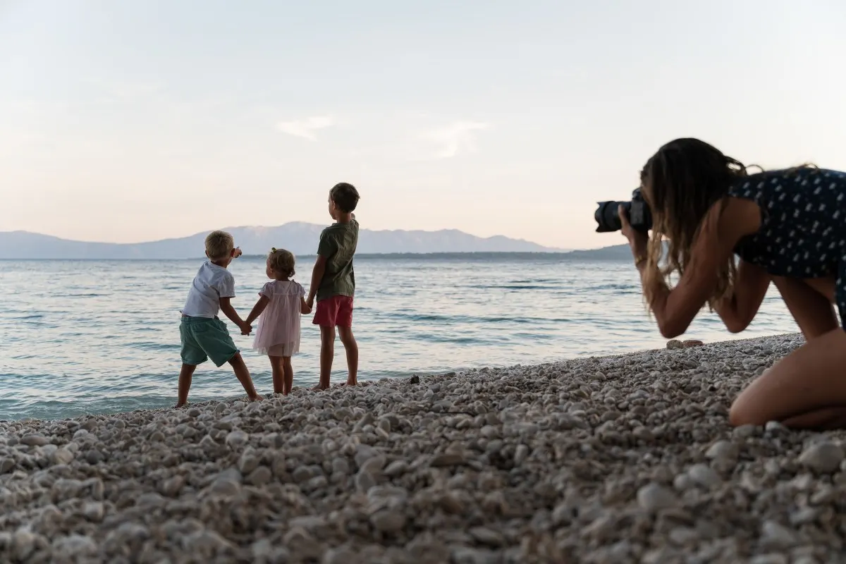 Trois frères et sœurs se tiennent sur une plage de galets et regardent l’eau, tandis que leur mère prend une photo d’eux en arrière-plan.