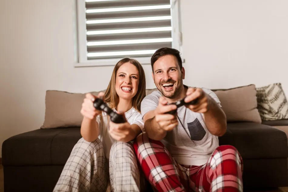 Deux personnes en pyjama jouent en riant avec des manettes de jeu dans le salon.