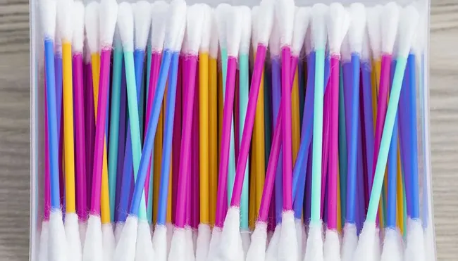 Contenitore pieno di cotton fioc colorati con bastoncini blu, viola, rosa, giallo e verde acqua e punte di cotone bianche.