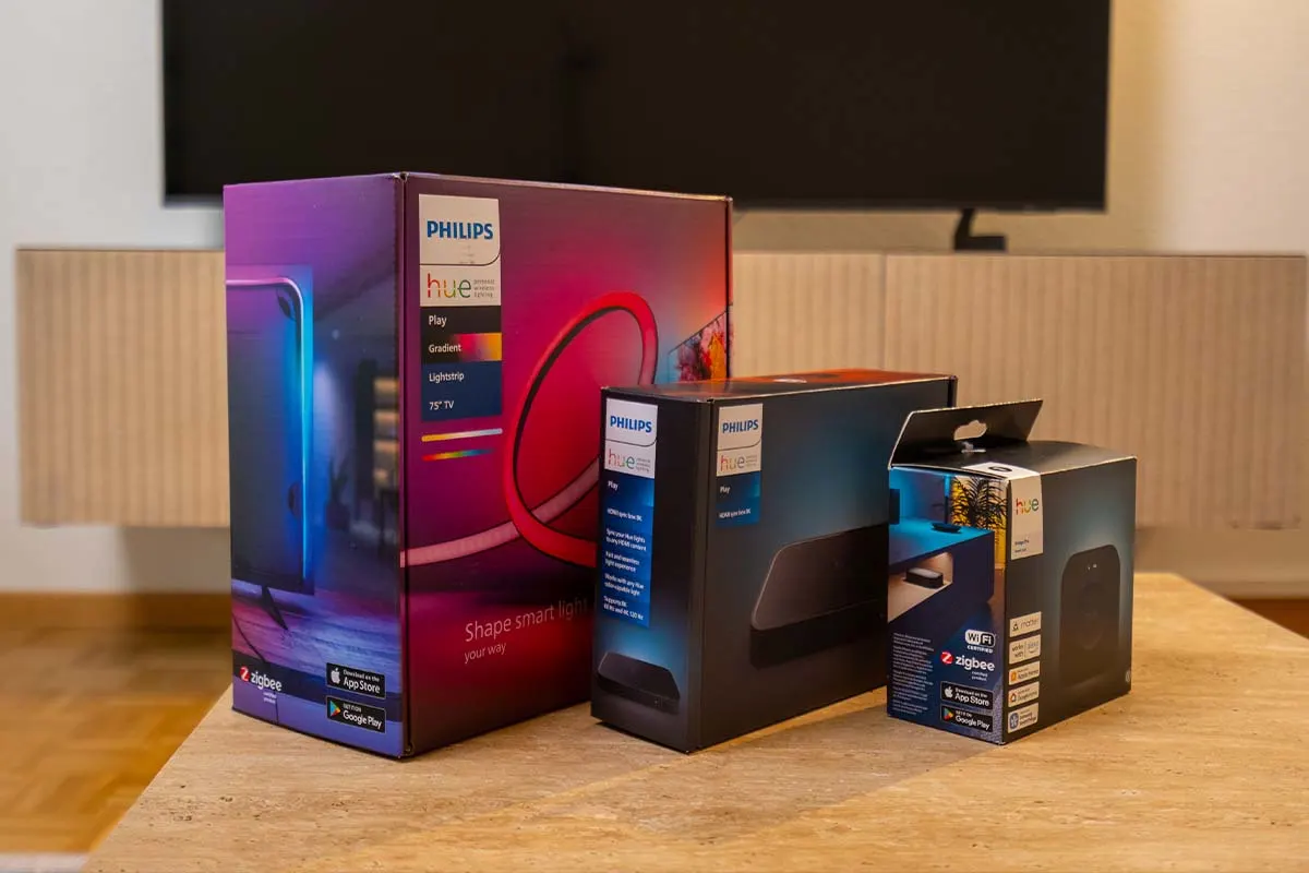 Mehrere Philips Hue Produkte stehen vor einem Fernseher auf einem Holztisch, darunter eine Hue Play Gradient Lightstrip Box und Zubehör für smarte Hintergrundbeleuchtung.