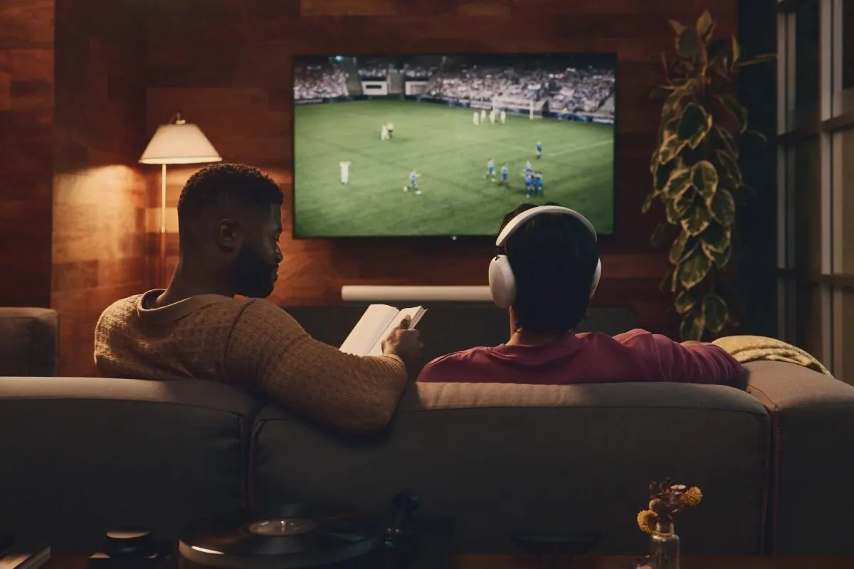 Due uomini sono seduti insieme sul divano: uno legge un libro mentre l’altro segue una partita di calcio in televisione.