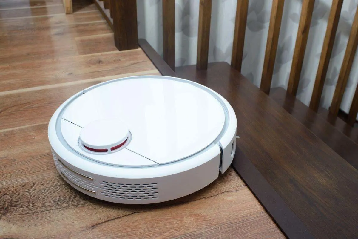 Robot aspirapolvere bianco su un pavimento in legno accanto a una scala.