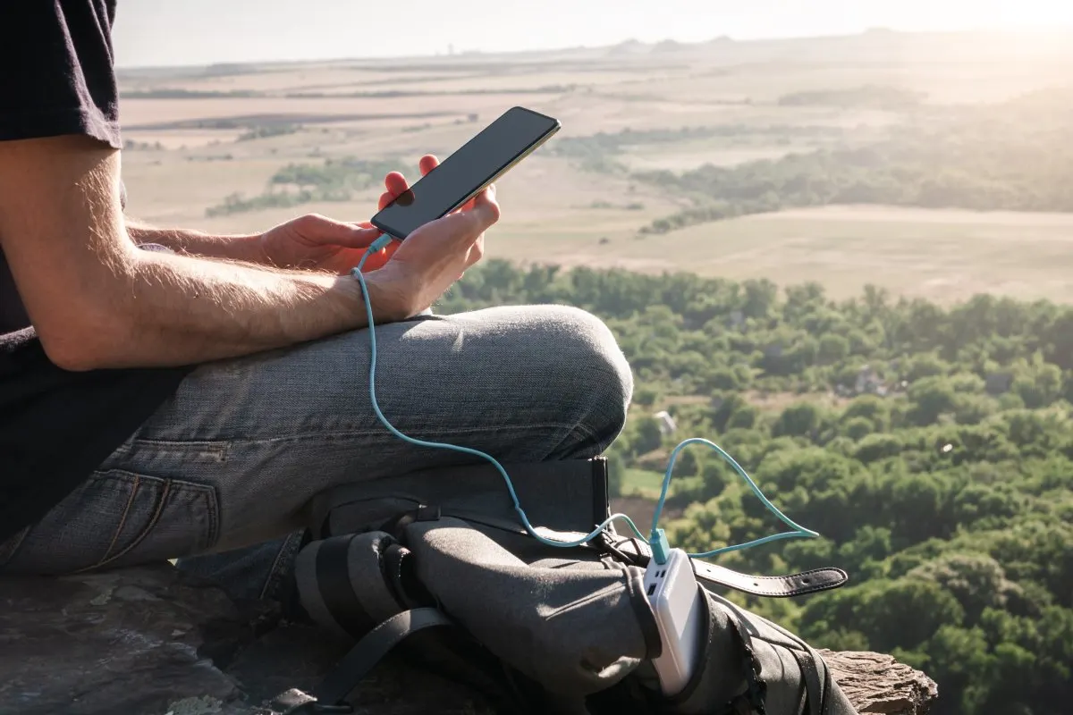 Una persona ricarica uno smartphone con una powerbank appoggiata su uno zaino, nella natura.