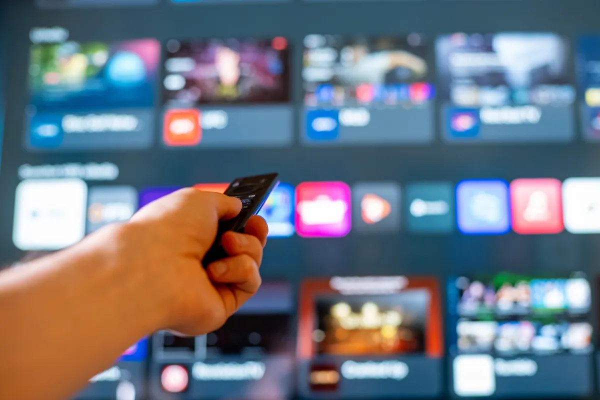 Main tenant une télécommande devant un smart TV avec un menu de streaming.