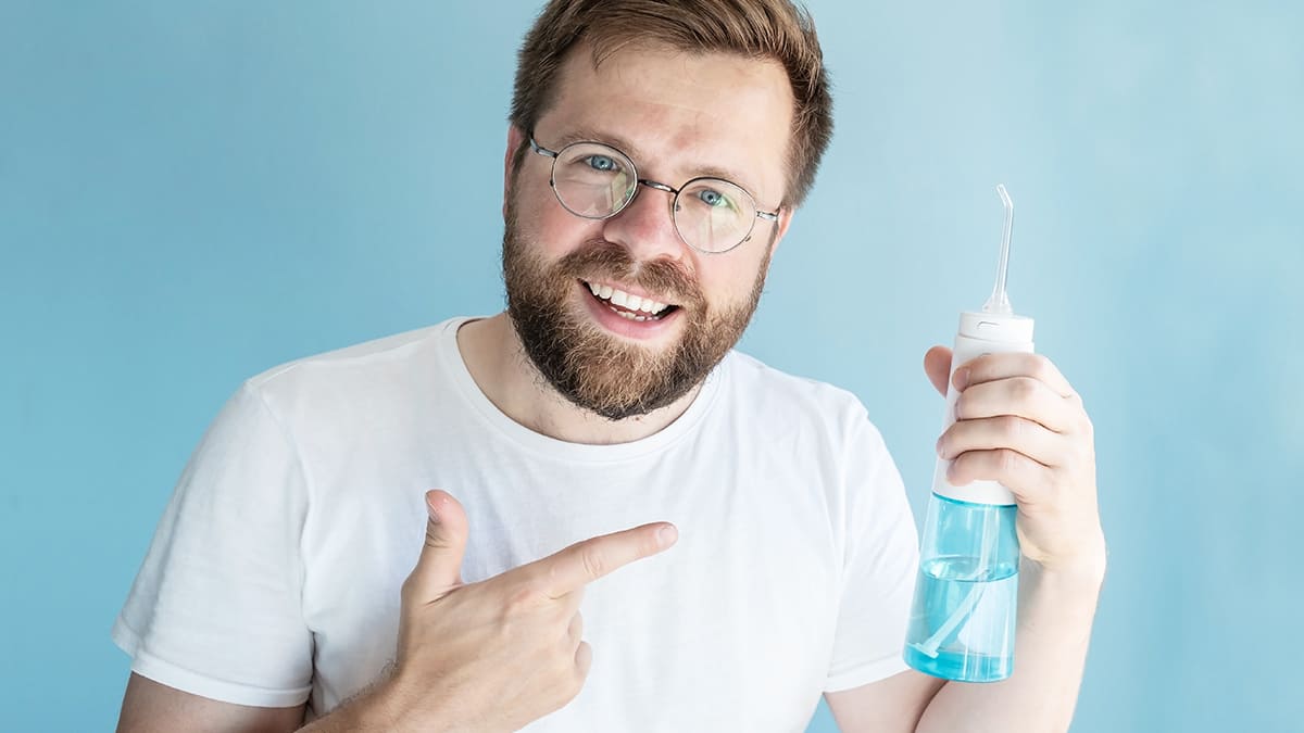 Uomo sorridente con occhiali che indica un idropulsore bianco e blu pieno d'acqua, su sfondo azzurro. Indossa una t-shirt bianca.