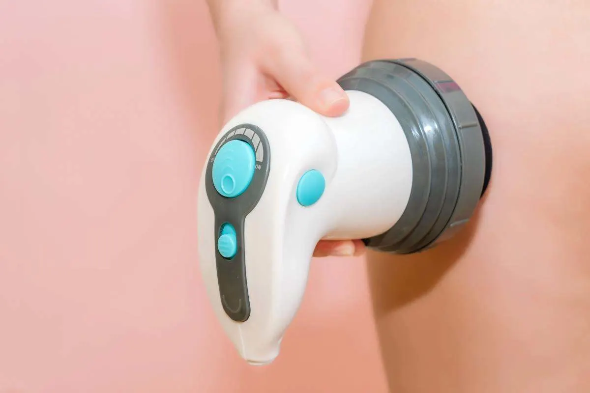 Mano che tiene un massaggiatore anticellulite elettrico con testina rotonda sulla pelle di una coscia, su sfondo rosa. 