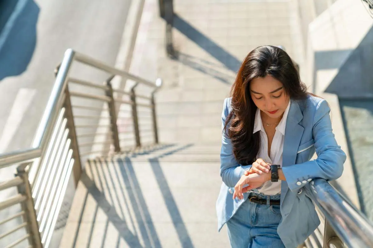 Femme d'affaires en blazer bleu clair et jean, appuyée contre une rambarde d'escalier en extérieur, le regard posé sur sa montre connectée.
