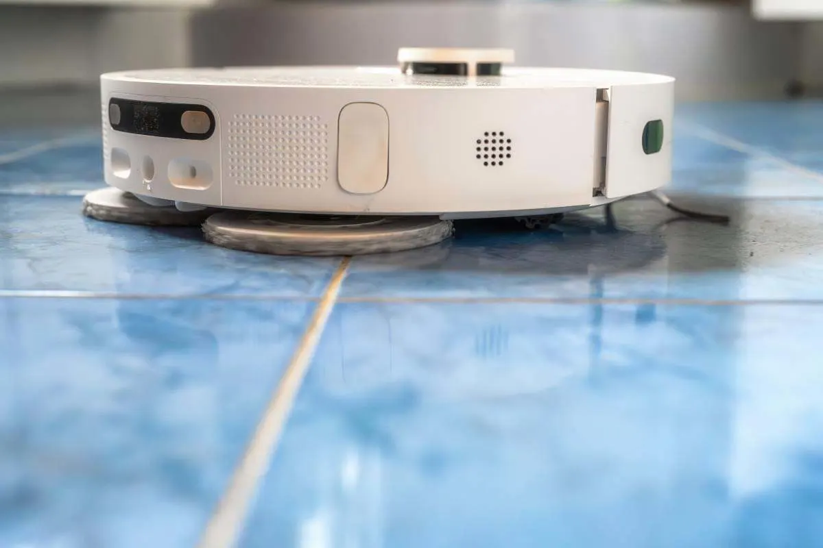 Gros plan sur un robot aspirateur laveur blanc à serpillières rotatives sur un carrelage bleu.